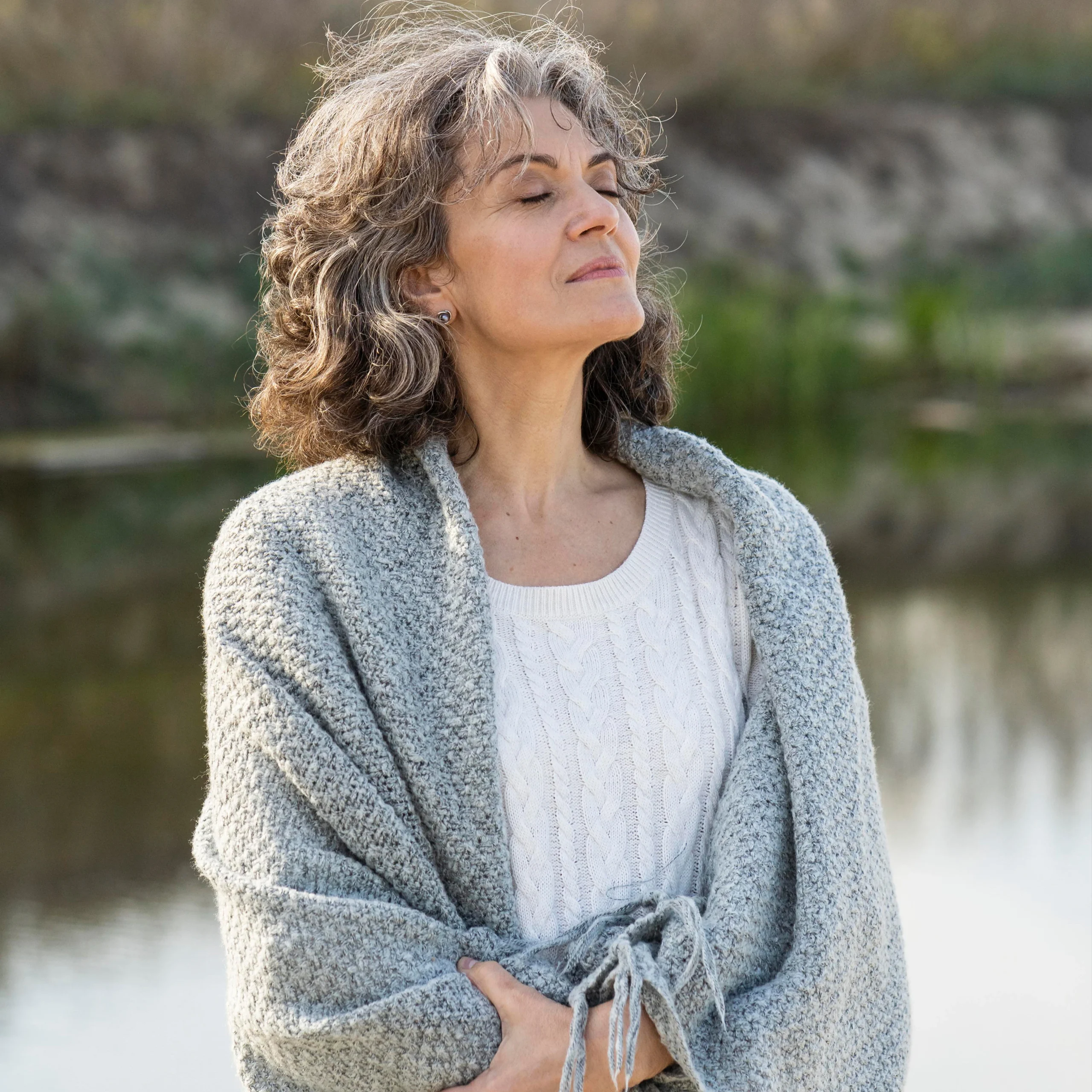 An older woman with gray curly hair stands outdoors by the water, eyes closed and smiling slightly. She is wrapped in a gray knit shawl over a white sweater, appearing peaceful and content.